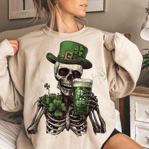 Op de afbeelding: Beige sweatshirt met een skelet dat een groene hoed met een klavertje draagt en een groen bier vasthoudt. Klavers omringen de borst van het skelet. Een leuk ontwerp voor St. Patrick's Day.