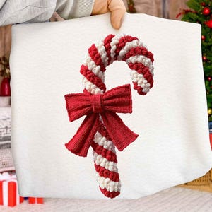 May include: A red and white striped candy cane with a red bow. The candy cane is made of a textured material and is a festive decoration. The bow is large and adds a decorative touch. The candy cane is a classic Christmas symbol.
