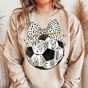 May include: A beige sweatshirt with a black and white polka dot bow on top of a black and white soccer ball with gold accents.