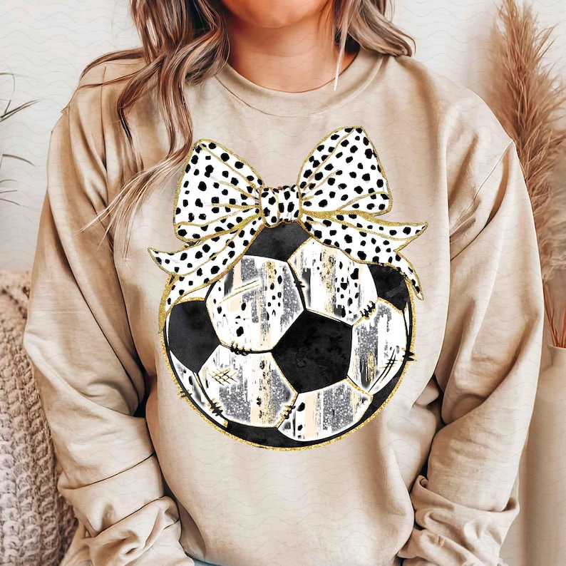 May include: A beige sweatshirt with a black and white polka dot bow on top of a black and white soccer ball with gold accents.