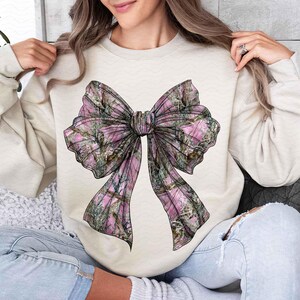 May include: A cream-colored sweatshirt featuring a large bow graphic. The bow has a pink and green camouflage pattern. The sweatshirt has long sleeves and a crew neck. The bow is the focal point of the design.