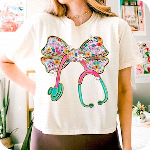 Peut inclure: T-shirt cr&egrave;me avec un n&oelig;ud floral color&eacute; et un motif de st&eacute;thoscope. Le n&oelig;ud est orn&eacute; de fleurs roses, bleues, jaunes et oranges. Le st&eacute;thoscope est rose et turquoise avec des d&eacute;tails dor&eacute;s, et de petites marguerites blanches pars&egrave;ment le motif.