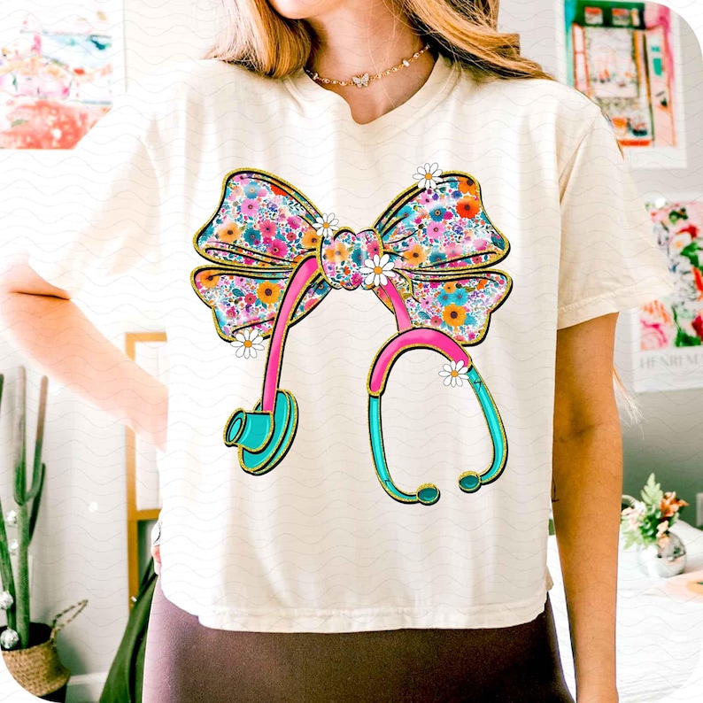 Peut inclure: T-shirt cr&egrave;me avec un n&oelig;ud floral color&eacute; et un motif de st&eacute;thoscope. Le n&oelig;ud est orn&eacute; de fleurs roses, bleues, jaunes et oranges. Le st&eacute;thoscope est rose et turquoise avec des d&eacute;tails dor&eacute;s, et de petites marguerites blanches pars&egrave;ment le motif.
