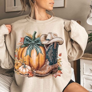 May include: A beige sweatshirt with a graphic print of a pumpkin, a cowboy boot, and a bow. The pumpkin is orange with green leaves and a green and white bow. The cowboy boot is brown with a blue and white design. The background is a light beige.
