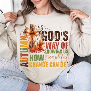 May include: A beige sweatshirt with a graphic design featuring a cross with fall leaves and the text "Autumn is God's way of showing us how beautiful change can be."