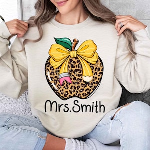 Puede incluir: Una sudadera beige con un dise&ntilde;o de manzana de estampado de leopardo. La manzana tiene un lazo amarillo y un l&aacute;piz que sobresale de ella. El texto "Mrs. Smith" est&aacute; impreso debajo de la manzana.