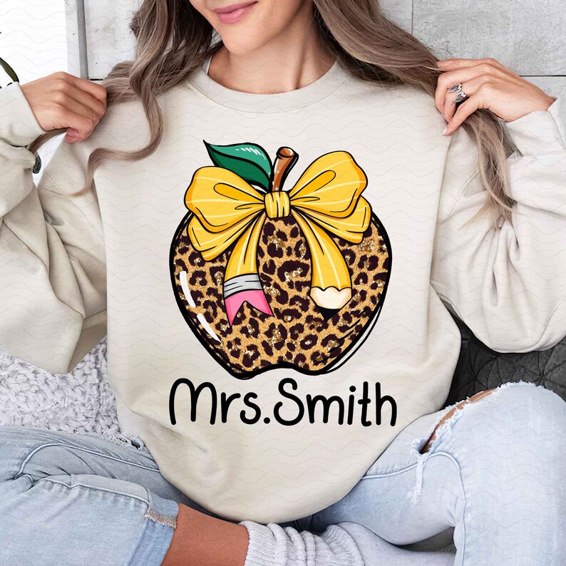 Puede incluir: Una sudadera beige con un dise&ntilde;o de manzana de estampado de leopardo. La manzana tiene un lazo amarillo y un l&aacute;piz que sobresale de ella. El texto "Mrs. Smith" est&aacute; impreso debajo de la manzana.