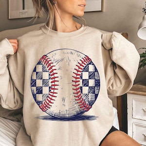 Puede incluir: Una sudadera beige con un gr&aacute;fico de una pelota de b&eacute;isbol con un patr&oacute;n a cuadros. La pelota de b&eacute;isbol est&aacute; dibujada en negro, rojo y blanco.