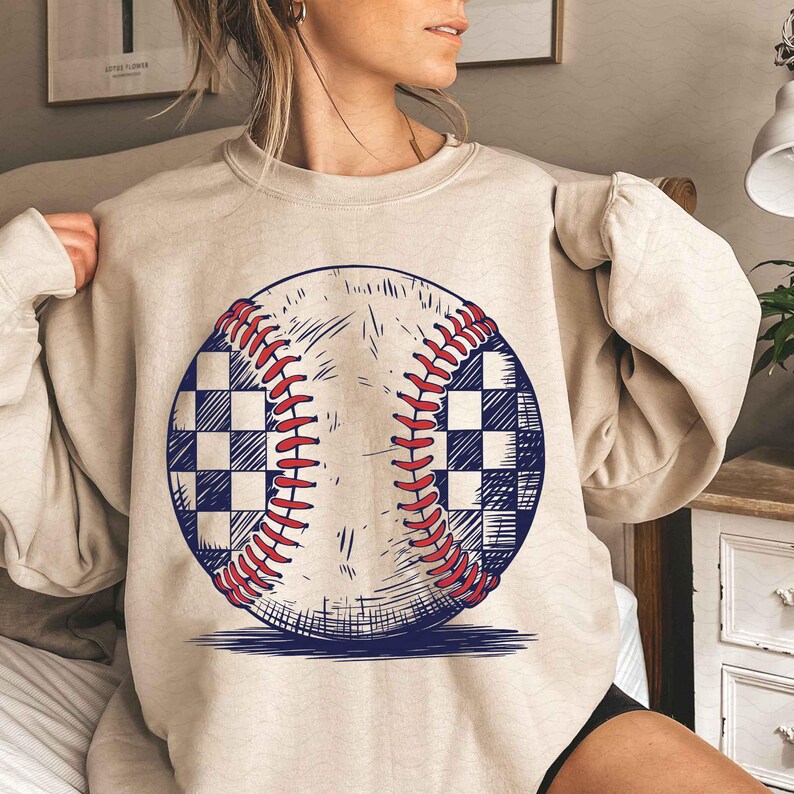 Puede incluir: Una sudadera beige con un gr&aacute;fico de una pelota de b&eacute;isbol con un patr&oacute;n a cuadros. La pelota de b&eacute;isbol est&aacute; dibujada en negro, rojo y blanco.
