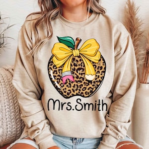 Puede incluir: Sudadera beige con un dise&ntilde;o de manzana de estampado de leopardo, un lazo amarillo y un l&aacute;piz. El texto "Mrs. Smith" est&aacute; impreso debajo de la manzana.