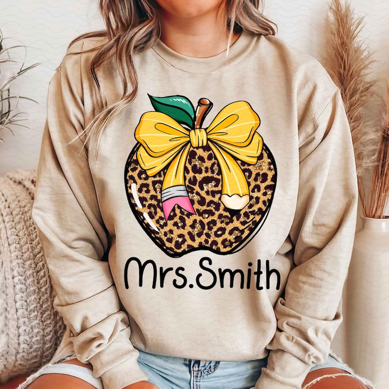 Puede incluir: Sudadera beige con un dise&ntilde;o de manzana de estampado de leopardo, un lazo amarillo y un l&aacute;piz. El texto "Mrs. Smith" est&aacute; impreso debajo de la manzana.