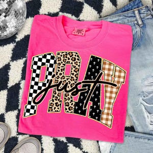 Könnte beinhalten: Ein leuchtend pinkfarbenes T-Shirt mit den Worten "PRAY just" in einer Mischung aus Mustern, darunter Schachbrettmuster, Leopardenmuster und Karomuster. Das T-Shirt ist gefaltet und liegt auf einer gemusterten Oberfläche mit Jeans und Turnschuhen.