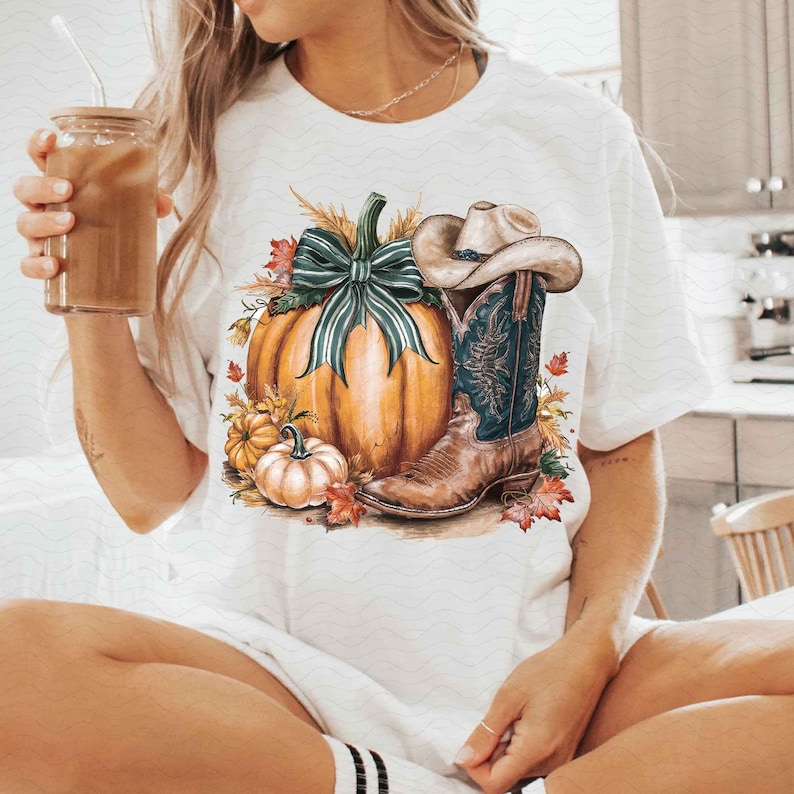 May include: White t-shirt with a graphic of a pumpkin, cowboy boot, and cowboy hat with a green bow. The pumpkin is orange and the boot is brown with blue accents.