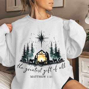 May include: White sweatshirt with a Christmas nativity scene graphic. The graphic features a star, a manger, a family, and animals. The text "the greatest gift of all" and "MATTHEW 1:21" is printed below the graphic.
