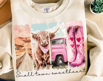 May include: A beige sweatshirt with a graphic design featuring a pink pickup truck, a highland cow, and pink cowboy boots. The graphic is surrounded by a distressed, watercolor-style border. The text "Small town sweetheart" is printed below the graphic.