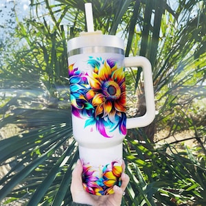 Pode incluir: Um copo branco grande com uma al&ccedil;a e uma palhinha. O copo &eacute; decorado com um design de girassol vibrante e colorido. O girassol tem p&eacute;talas amarelas, laranja, azuis e roxas. O fundo est&aacute; desfocado com folhagem verde.