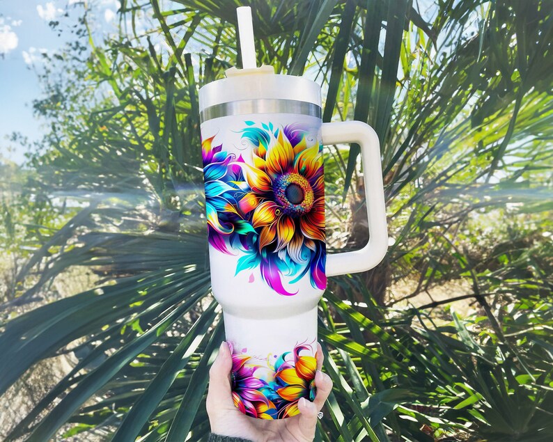 Pode incluir: Um copo branco grande com uma al&ccedil;a e uma palhinha. O copo &eacute; decorado com um design de girassol vibrante e colorido. O girassol tem p&eacute;talas amarelas, laranja, azuis e roxas. O fundo est&aacute; desfocado com folhagem verde.