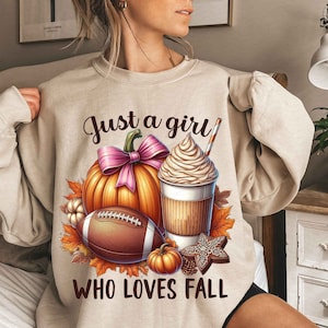 Puede incluir: Una sudadera beige con un estampado gráfico de una calabaza, una pelota de fútbol, una taza de café con crema batida y un lazo rosa. El texto "Just a girl who loves fall" está impreso en la sudadera.