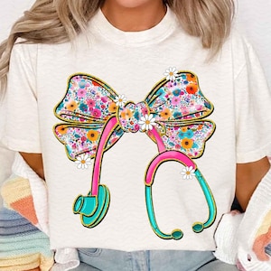 Peut inclure: T-shirt cr&egrave;me avec un n&oelig;ud floral et un motif de st&eacute;thoscope. Le n&oelig;ud pr&eacute;sente un motif floral color&eacute;, et le st&eacute;thoscope est rose et turquoise avec des accents dor&eacute;s. De petits accents de marguerites blanches sont plac&eacute;s autour du motif.