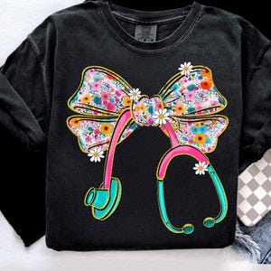 Peut inclure: Sweat-shirt noir avec un n&oelig;ud floral et un motif de st&eacute;thoscope. Le n&oelig;ud est orn&eacute; de fleurs roses, oranges et bleues. Le st&eacute;thoscope est rose et turquoise, avec des accents de marguerites, cr&eacute;ant un design sur le th&egrave;me m&eacute;dical.