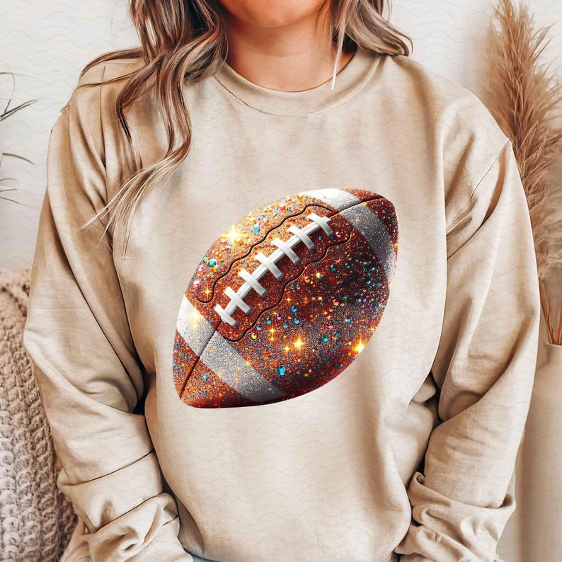May include: A beige sweatshirt with a glittery brown football design. The football has white stitching and a white stripe around the middle.