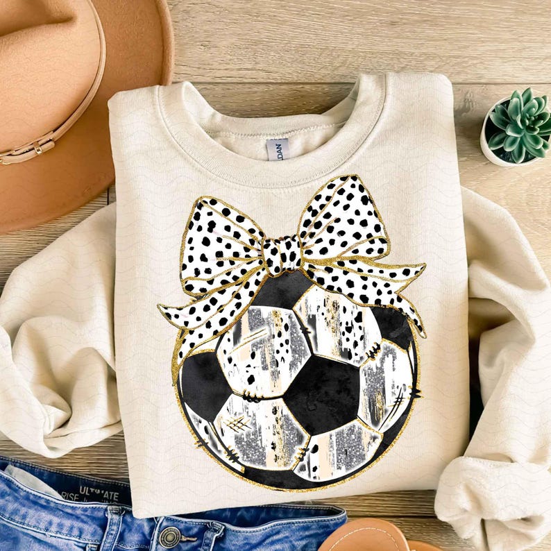 May include: A cream-colored sweatshirt with a black and white polka dot bow on top of a black and white soccer ball.
