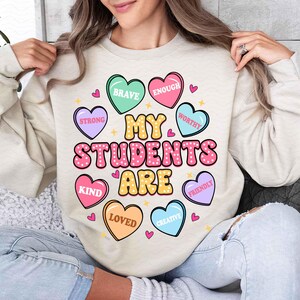 May include: A light beige sweatshirt with the text "My Students Are" in pink and yellow letters surrounded by colorful candy hearts with words like "Brave", "Enough", "Strong", "Worthy", "Kind", "Loved", "Creative", and "Friendly".