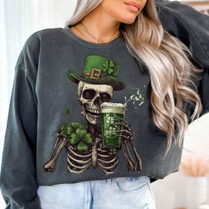 Peut inclure: Sweat-shirt gris fonc&eacute; avec un squelette portant un chapeau vert avec un tr&egrave;fle et tenant une bi&egrave;re verte. Le squelette a un bouquet de tr&egrave;fles et le verre &agrave; bi&egrave;re a un motif de tr&egrave;fle. Le sweat-shirt est sur le th&egrave;me de la Saint-Patrick.