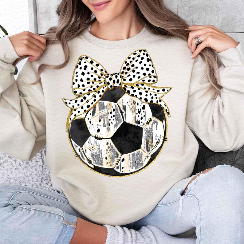 May include: A cream-colored sweatshirt with a black and white polka dot bow on top of a black and white soccer ball with gold accents.