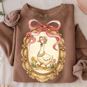 以下が含まれることがあります： 帽子とネックレスを身に着けたガチョウのイラストが額縁に入った、茶色のスウェットシャツ。フレームの上部にはピンクのリボンの弓が付いています。スウェットシャツは折りたたまれており、袖とネックラインが見えます。