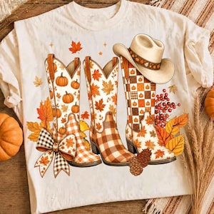 Puede incluir: Una camiseta de color crema con un estampado gráfico de dos botas de vaquero y un sombrero de vaquero. Las botas están decoradas con hojas de otoño naranjas, calabazas y un patrón de cuadros vichy marrón. Incluye un lazo grande, piñas y bayas.