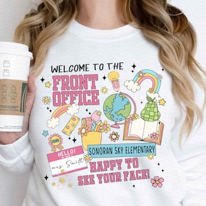 May include: White sweatshirt with a pink and blue graphic design that says "Welcome to the Front Office". The graphic includes a rainbow, a globe, a book, a pencil, a pear, and a flower pot with school supplies. The text "Sonoran Sky Elementary" and "Happy to See Your Face!" are also included in the design.