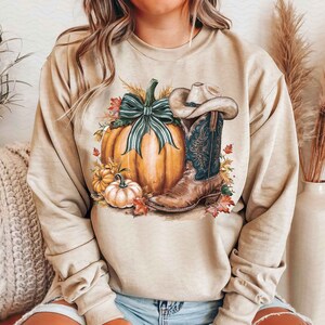 May include: A beige sweatshirt with a graphic print of a pumpkin, a cowboy boot, and fall leaves. The pumpkin has a green ribbon and the boot has a brown hat.