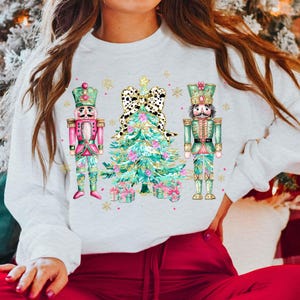 May include: White sweatshirt with a Christmas-themed design featuring two nutcracker soldiers flanking a green Christmas tree with a pink bow. The tree is decorated with gold glitter and pink ornaments.