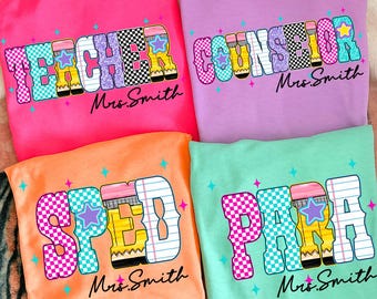 May include: Four brightly coloured t-shirts with school-themed designs. Each shirt displays a different word: "TEACHER", "COUNSELOR", "SPED", and "PARA", with a star and pencil graphic. The name "Mrs. Smith" is printed below each word.