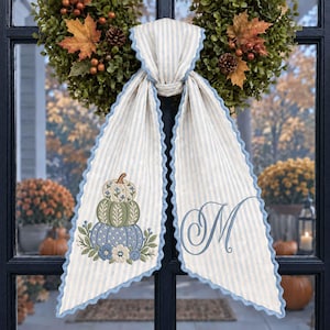 May include: A light blue and white striped fabric door hanger featuring a scalloped edge. The left side displays an embroidered stack of three pumpkins with floral accents, while the right side features an embroidered cursive letter M.