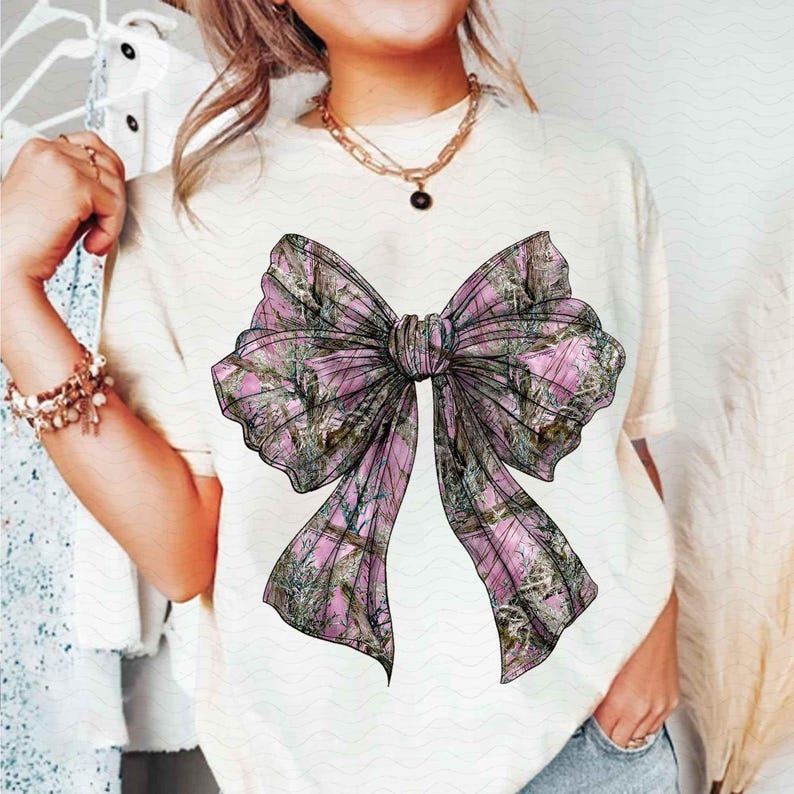 May include: Cream-colored t-shirt featuring a large bow graphic. The bow has a pink and green camouflage pattern. The shirt is a casual style, suitable for everyday wear.