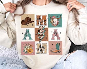 Puede incluir: Una sudadera de color crema con un diseño gráfico de un sombrero de vaquero, un rayo, una bota y las letras "MAMA" en un estilo vintage.