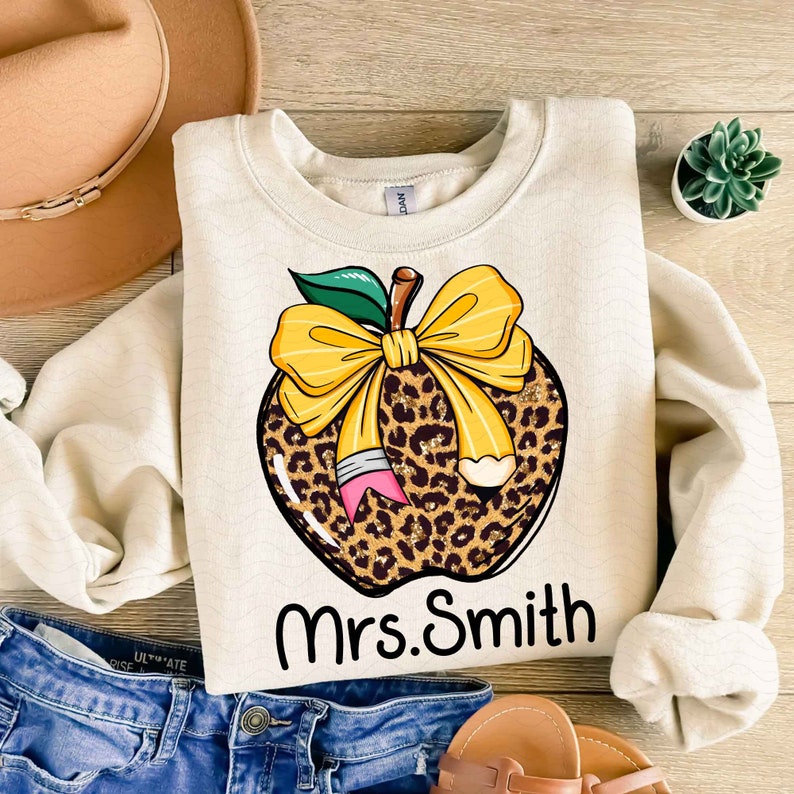 Puede incluir: Una sudadera beige con un dise&ntilde;o de manzana de estampado de leopardo. La manzana tiene un lazo amarillo y un l&aacute;piz que sobresale de ella. El texto "Mrs. Smith" est&aacute; impreso debajo de la manzana.