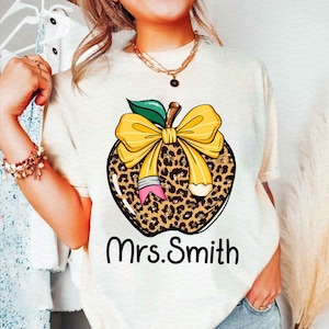 Puede incluir: Camiseta blanca con un dise&ntilde;o de manzana de estampado de leopardo. La manzana tiene un lazo amarillo y un l&aacute;piz que sobresale. El texto "Mrs. Smith" est&aacute; impreso debajo de la manzana.