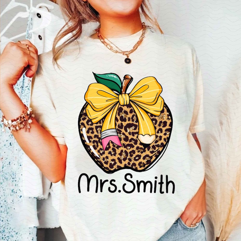 Puede incluir: Camiseta blanca con un dise&ntilde;o de manzana de estampado de leopardo. La manzana tiene un lazo amarillo y un l&aacute;piz que sobresale. El texto "Mrs. Smith" est&aacute; impreso debajo de la manzana.