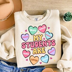 May include: A beige sweatshirt with the text "My Students Are" in pink and white letters.  Surrounding the text are pink, blue, green, and yellow heart-shaped designs with words like "Brave", "Strong", "Kind", "Loved", "Creative", "Enough", "Worthy", and "Friendly".