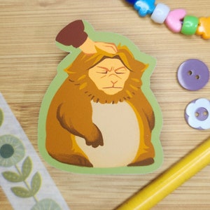 May include: A sticker of a brown monkey with a green background. The monkey has a grumpy expression and is being petted on the head by a hand.