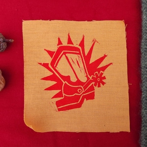 May include: A square, tan fabric patch with a red graphic of a gas mask and a starburst design. The patch is set against a red background.