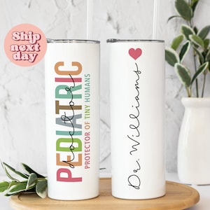 May include: Two white stainless steel tumblers with lids and straws. One tumbler has the text "PEDIATRIC" in rainbow colors with the words "protector of tiny humans" below. The other tumbler has the text "Dr. Williams" in black cursive with a small pink heart above the text.