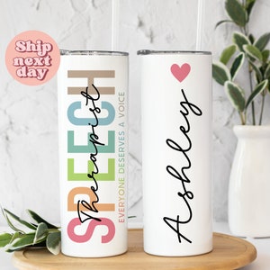 May include: Two white stainless steel tumblers with colorful rainbow designs. One tumbler says "Speech Therapist" with the text "Everyone deserves a voice" in smaller font. The other tumbler has the name "Ashley" written in cursive with a pink heart above it.