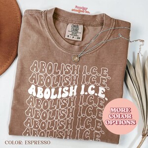 May include: Espresso-colored t-shirt with the text "ABOLISH I.C.E." in a retro font. The shirt has a crew neck and short sleeves. A silver necklace with a pendant is draped across the shirt. The words "MORE COLOR OPTIONS" are in a pink circle.