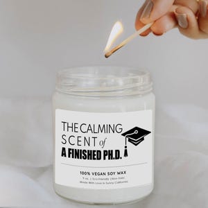 A hand lighting a soy wax candle labeled "The calming scent of a finished PhD."