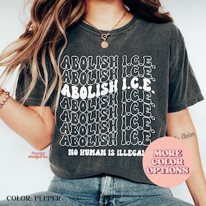 May include: Pepper-colored t-shirt with the text "ABOLISH I.C.E." repeated in a wavy, white font. The phrase "NO HUMAN IS ILLEGAL" is printed below the text. A pink circle with "MORE COLOR OPTIONS" is in the lower right.