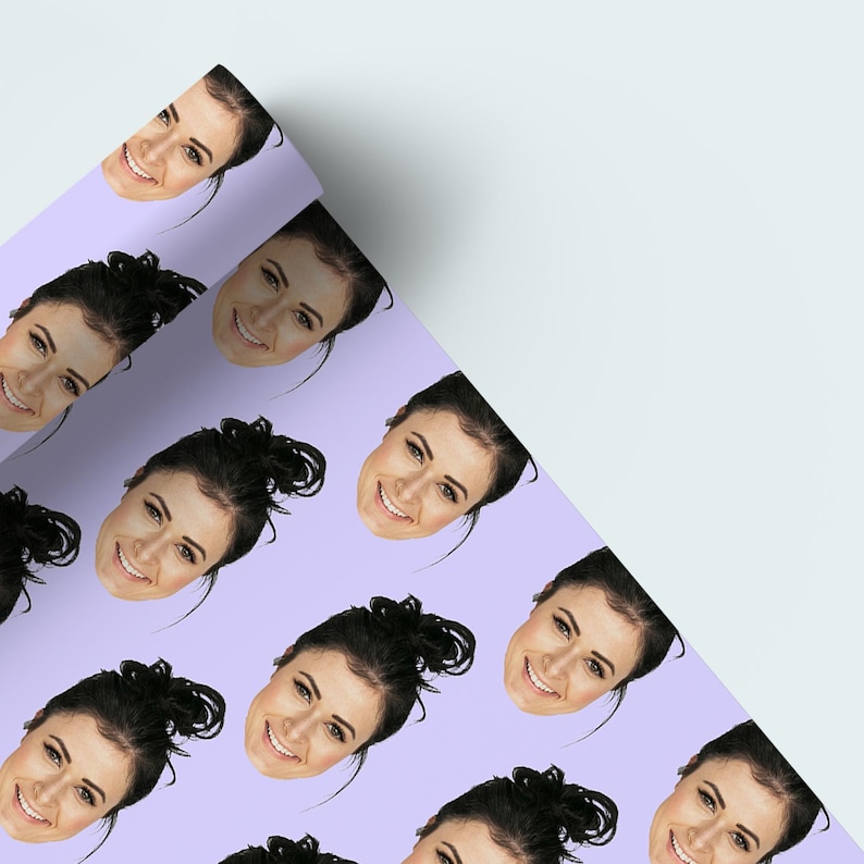 May include: A roll of wrapping paper featuring a repeating pattern of a woman's face with dark hair and a smile. The background is a light purple color. The paper is partially unrolled, revealing a white surface.